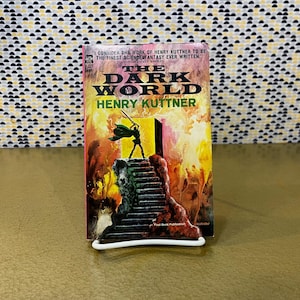 Op de afbeelding: Een paperback boek getiteld "The Dark World" van Henry Kuttner, met een levendige omslagillustratie. De omslag toont een figuur met een zwaard op een trap, tegen een vurige achtergrond. Het boek wordt op een witte standaard gepresenteerd.
