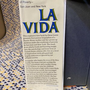 Op de afbeelding: Een boekomslag met de titel "La Vida" in blauwe en gouden letters. Het boek gaat over een Puerto Ricaans gezin dat in armoede leeft in San Juan en New York. De tekst op de omslag beschrijft het boek als een explosief nieuw boek van Oscar Lewis, dat persoonlijke biografie&euml;n presenteert van een Puerto Ricaanse moeder en haar volwassen kinderen onder de omstandigheden van armoede.