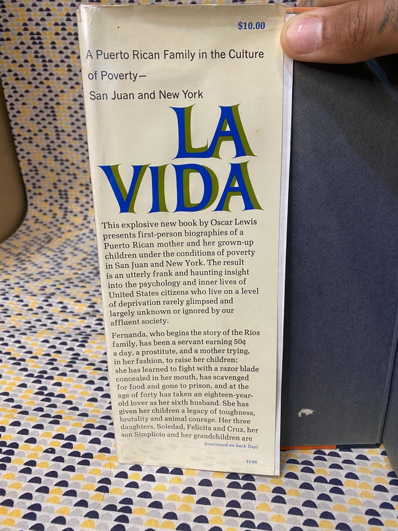Op de afbeelding: Een boekomslag met de titel "La Vida" in blauwe en gouden letters. Het boek gaat over een Puerto Ricaans gezin dat in armoede leeft in San Juan en New York. De tekst op de omslag beschrijft het boek als een explosief nieuw boek van Oscar Lewis, dat persoonlijke biografie&euml;n presenteert van een Puerto Ricaanse moeder en haar volwassen kinderen onder de omstandigheden van armoede.
