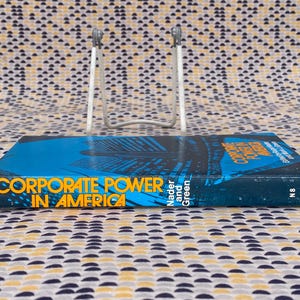 May include: A blue and yellow paperback book titled "Corporate Power in America" by Nader and Green. The book is on a book stand and the cover features a cityscape.