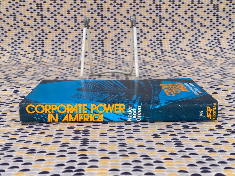 May include: A blue and yellow paperback book titled "Corporate Power in America" by Nader and Green. The book is on a book stand and the cover features a cityscape.