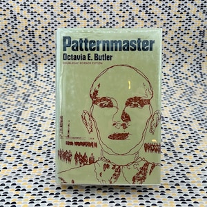 May include: A paperback copy of "Patternmaster" by Octavia E. Butler. The cover displays a brown illustration of a person's face and a crowd on a light green backdrop. The book is part of the Doubleday Science Fiction series.