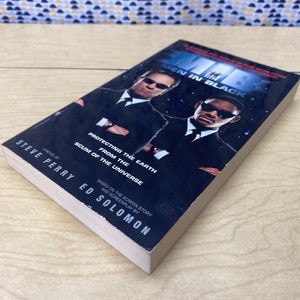 Puede incluir: Una portada de libro de bolsillo para la novela "Men in Black" de Steve Perry. La portada presenta a dos hombres con trajes negros, uno con una expresi&oacute;n seria y el otro con una sonrisa burlona. El texto de la portada dice "Protegiendo la Tierra del desecho del universo".