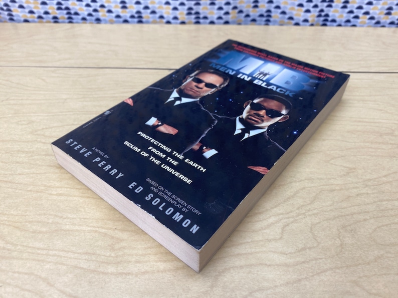 Puede incluir: Una portada de libro de bolsillo para la novela "Men in Black" de Steve Perry. La portada presenta a dos hombres con trajes negros, uno con una expresi&oacute;n seria y el otro con una sonrisa burlona. El texto de la portada dice "Protegiendo la Tierra del desecho del universo".