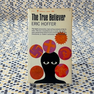 Puede incluir: Libro de bolsillo vintage titulado "The True Believer" de Eric Hoffer. La portada presenta una silueta negra con diseños circulares y el título en negrita. El libro está sobre un fondo estampado.