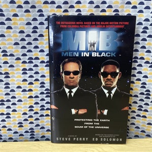 Puede incluir: Una portada de libro de bolsillo para la novela "Men in Black" basada en la pel&iacute;cula. La portada presenta una foto en blanco y negro de los dos personajes principales, Tommy Lee Jones y Will Smith, vistiendo trajes negros y gafas de sol. El t&iacute;tulo "Men in Black" est&aacute; en letras blancas grandes sobre un fondo negro. El texto "Protecting the Earth from the Scum of the Universe" est&aacute; debajo del t&iacute;tulo. Los nombres de los autores, Steve Perry y Ed Solomon, est&aacute;n en la parte inferior de la portada.