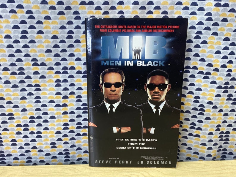 Puede incluir: Una portada de libro de bolsillo para la novela "Men in Black" basada en la pel&iacute;cula. La portada presenta una foto en blanco y negro de los dos personajes principales, Tommy Lee Jones y Will Smith, vistiendo trajes negros y gafas de sol. El t&iacute;tulo "Men in Black" est&aacute; en letras blancas grandes sobre un fondo negro. El texto "Protecting the Earth from the Scum of the Universe" est&aacute; debajo del t&iacute;tulo. Los nombres de los autores, Steve Perry y Ed Solomon, est&aacute;n en la parte inferior de la portada.