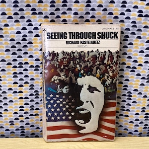 May include: A vintage paperback book titled "Seeing Through Shuck" by Richard Kostelanetz, Editor. The cover features a black and white illustration of a person's face with an open mouth, superimposed on an American flag.