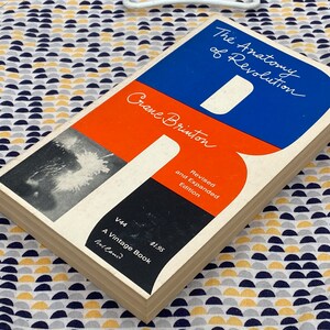 Puede incluir: Un libro de bolsillo vintage titulado "The Anatomy of Revolution" de Crane Brinton. La portada presenta un dise&ntilde;o rojo, azul y negro con el t&iacute;tulo en texto blanco. El libro es una edici&oacute;n revisada y ampliada y forma parte de la serie Vintage Book.