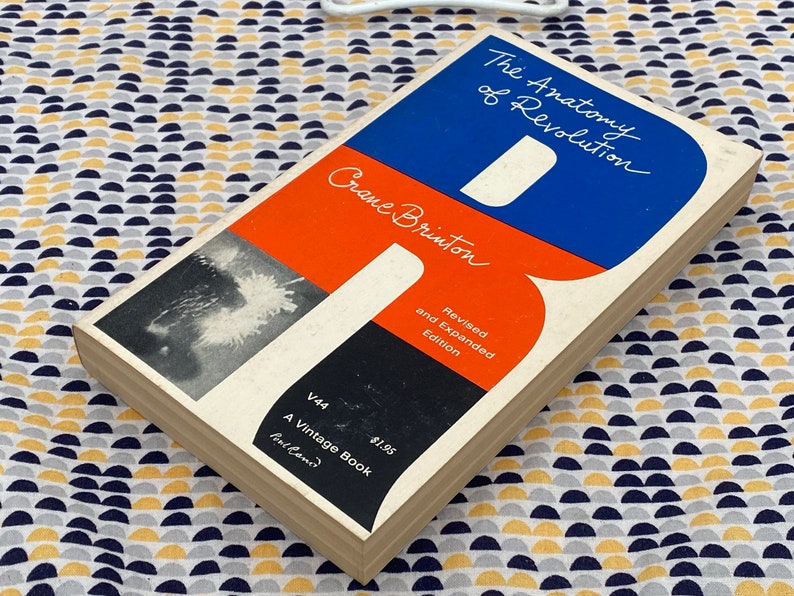 Puede incluir: Un libro de bolsillo vintage titulado "The Anatomy of Revolution" de Crane Brinton. La portada presenta un dise&ntilde;o rojo, azul y negro con el t&iacute;tulo en texto blanco. El libro es una edici&oacute;n revisada y ampliada y forma parte de la serie Vintage Book.