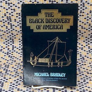May include: A hardcover book titled "The Black Discovery of America" by Michael Bradley. The cover is dark blue with gold lettering and an illustration of a ship. The book explores evidence of voyages by West African mariners.