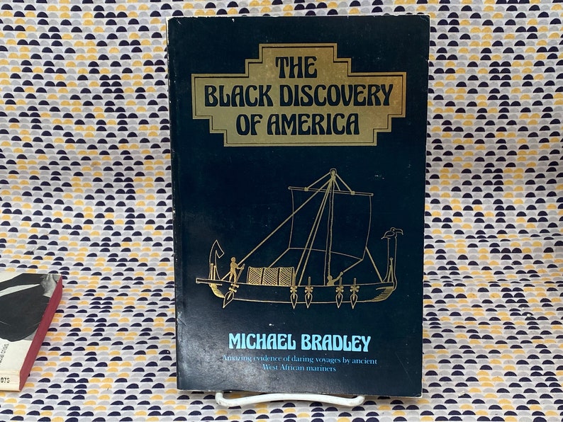 May include: A hardcover book titled "The Black Discovery of America" by Michael Bradley. The cover is dark blue with gold lettering and an illustration of a ship. The book explores evidence of voyages by West African mariners.