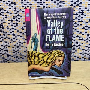 Op de afbeelding: Een vintage paperback boek getiteld "Valley of the Flame" van Henry Kuttner. De omslag toont een vrouw met blond haar en een futuristisch figuur. Het boek staat op een witte metalen standaard.