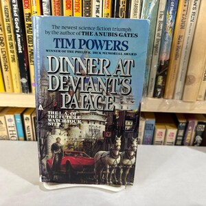 May include: A paperback book titled "Dinner at Deviant's Palace" by Tim Powers. The cover features a man, a red car, and two white horses. The book is set against a backdrop of other books on a shelf.