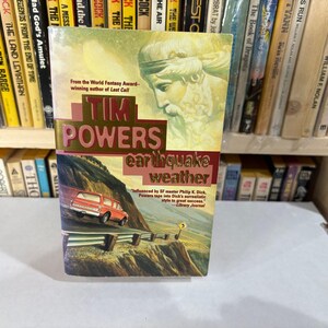 May include: A paperback book titled "Earthquake Weather" by Tim Powers. The cover features a red vehicle driving on a mountain road, with the author's name in large, gold-colored letters. The background includes a depiction of a classical figure.
