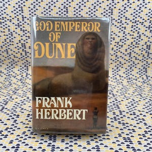 May include: A hardcover book titled "God Emperor of Dune" by Frank Herbert. The book cover features a sphinx-like figure in shades of brown and tan against a desert landscape. The title and author's name are in bold, white lettering.