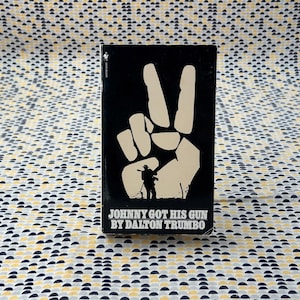 May include: A paperback book titled "Johnny Got His Gun" by Dalton Trumbo. The cover is black with a large, tan hand making a peace sign. Below the hand is a silhouette of a person holding a rifle.