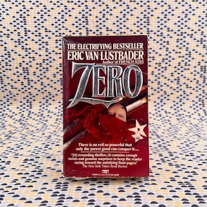 Puede incluir: Un libro de tapa dura titulado "ZERO" de Eric Van Lustbader, con una cubierta burdeos y letras plateadas. La portada presenta una persona con una prenda roja y una estrella blanca. El libro es un thriller, según The New York Times Book Review.