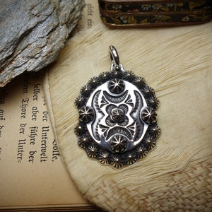 May include: Silver pendant with an oval design and intricate starburst detailing. The pendant has a bail for a chain and is set against a textured, off-white surface. The pendant's design includes a central floral motif.