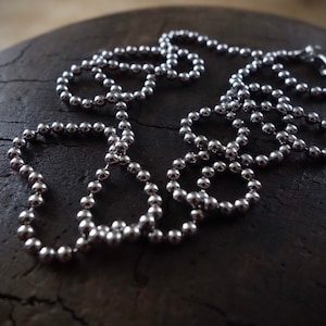 May include: A silver chain necklace made of small, round beads. The chain is lying on a dark, textured surface.
