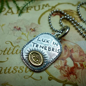 May include: Silver pendant necklace with the inscription "Lux in Tenebris". The pendant features a sunburst design with an eye in the center, and is on a ball chain necklace. The pendant is on a vintage book.