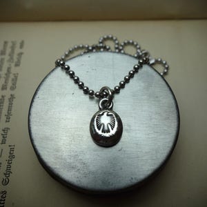 Könnte beinhalten: Ein silberfarbener Metallanhänger mit einem geprägten Adlerdesign auf einer ovalen Oberfläche, der an einer passenden silberfarbenen Kugelkette hängt. Der Anhänger ist auf einer runden Metallbasis zentriert und zeigt ein poliertes Finish.