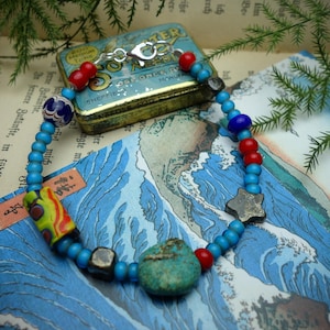 May include: A handmade beaded bracelet with turquoise, red, blue, and silver beads. The bracelet features a large turquoise stone, a star-shaped bead, and a colorful cylindrical bead. A small, vintage tin is in the background.