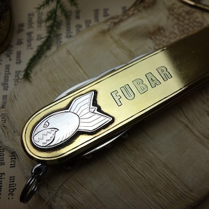 Pode incluir: Uma multi-ferramenta dourada com a palavra "FUBAR" gravada na lateral e um emblema em forma de bomba. A ferramenta é exibida em um fundo de estilo vintage, sugerindo um tema militar ou de novidade.