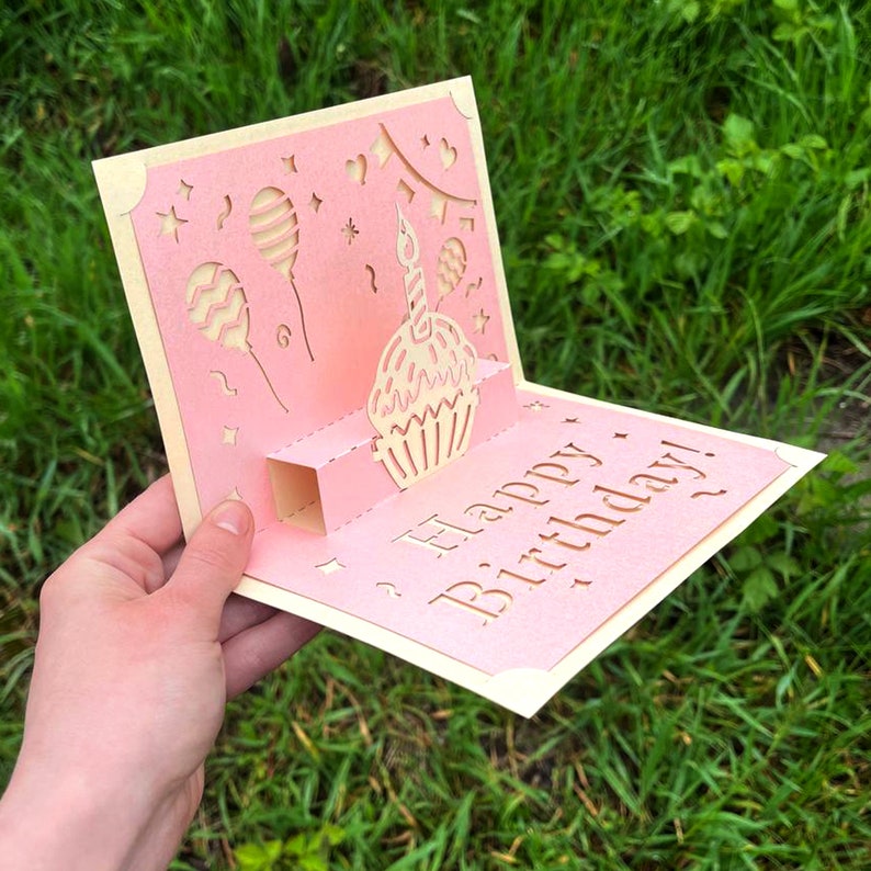 Closer handheld view of the dimensional 3D cupcake pop-up card template, emphasizing the precise laser-cut patterns for the central cupcake, balloons, and "Happy Birthday!" text against a natural blurry background.