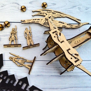 May include: A wooden dragon model kit with a dragon, two figures, and a catapult. The dragon is in a flying pose with its wings spread. The figures are standing on wooden bases. The catapult is made of wood and has a string attached to it. The kit is on a white wooden surface.
