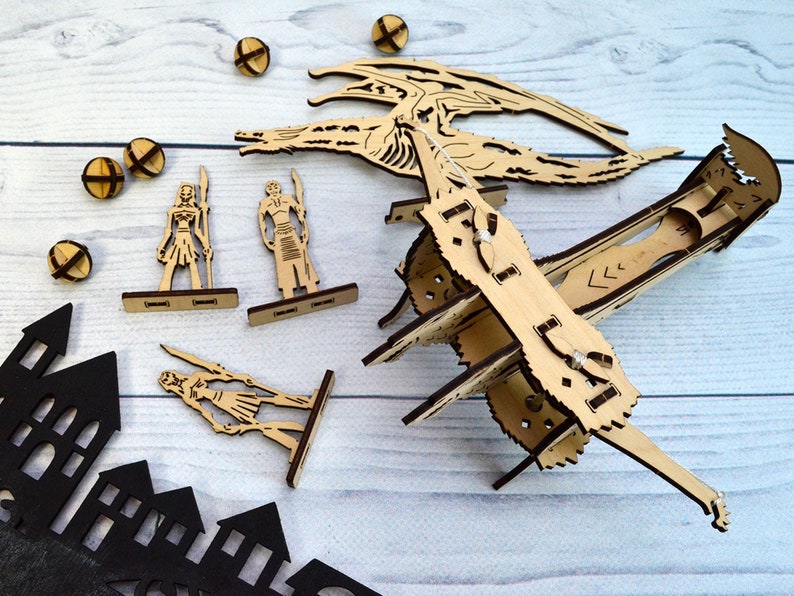 May include: A wooden dragon model kit with a dragon, two figures, and a catapult. The dragon is in a flying pose with its wings spread. The figures are standing on wooden bases. The catapult is made of wood and has a string attached to it. The kit is on a white wooden surface.
