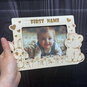 May include: A wooden photo frame with a cutout of a bunny and a bear. The frame has a space for a photo and the words "FIRST NAME" at the top.