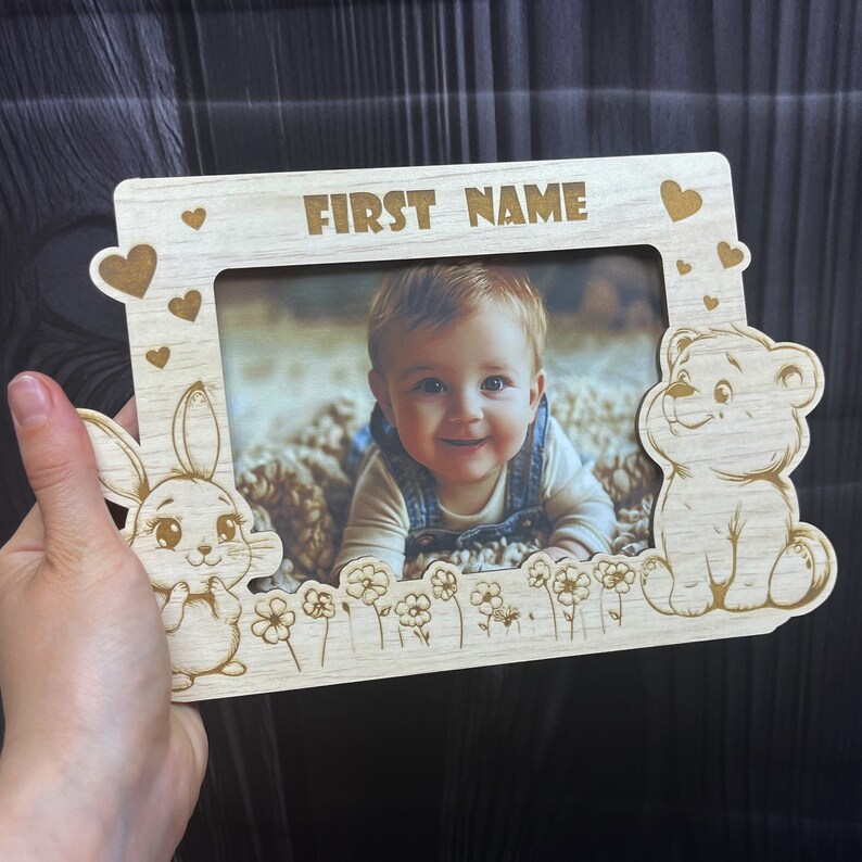 May include: A wooden photo frame with a cutout of a bunny and a bear. The frame has a space for a photo and the words "FIRST NAME" at the top.