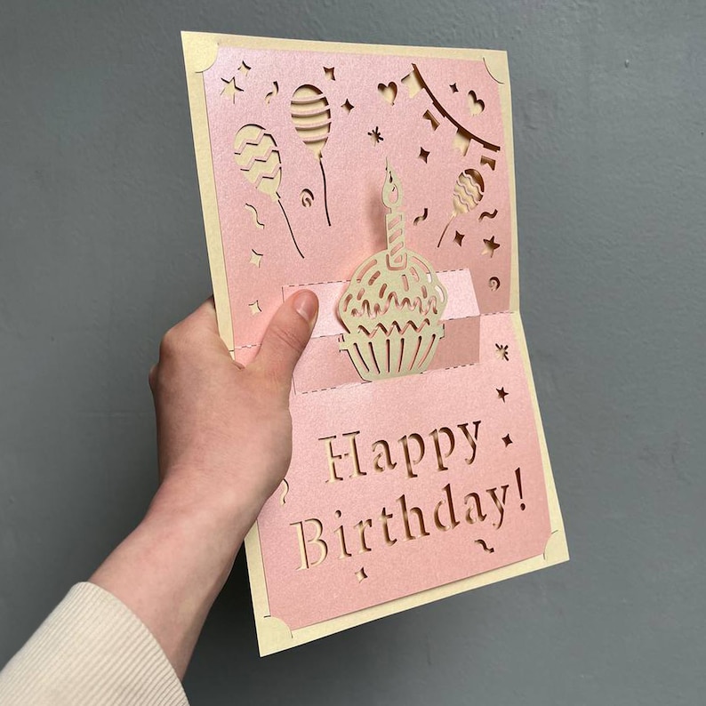 Handheld view of a 3D cupcake pop up card template. Demonstrates the intricate laser cut details and folding mechanism for a professional handmade DIY gift.
