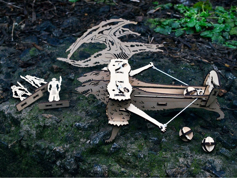 May include: A wooden model of a dragon with three heads, a crossbow, and four figures. The dragon is made of laser-cut wood and has a detailed design. The crossbow is also made of wood and has a string attached to it. The figures are small and are also made of wood.