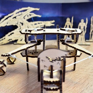 May include: A wooden toy catapult with a white string and four wooden balls. The catapult is made of wood and has a brown finish. The catapult is on a wooden table.