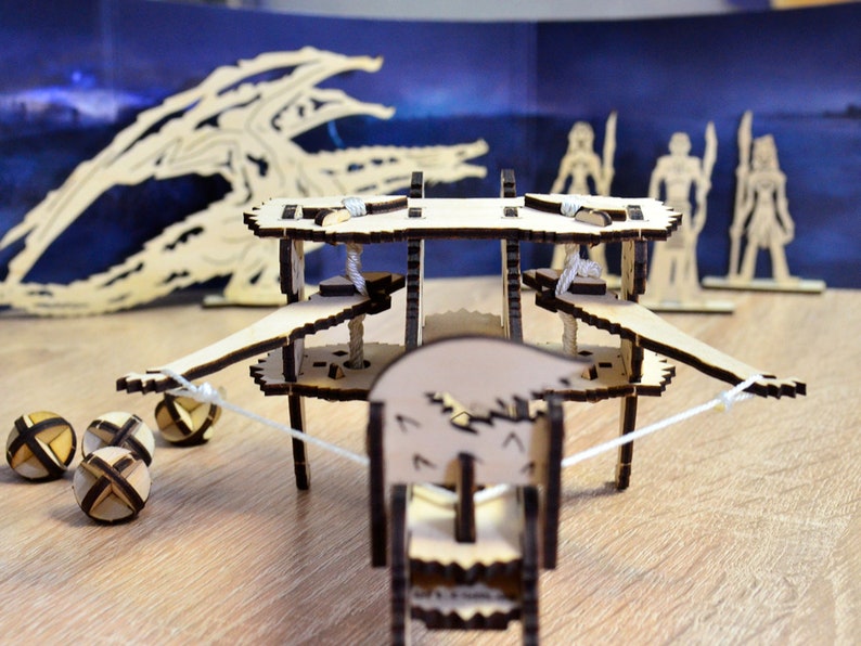 May include: A wooden toy catapult with a white string and four wooden balls. The catapult is made of wood and has a brown finish. The catapult is on a wooden table.
