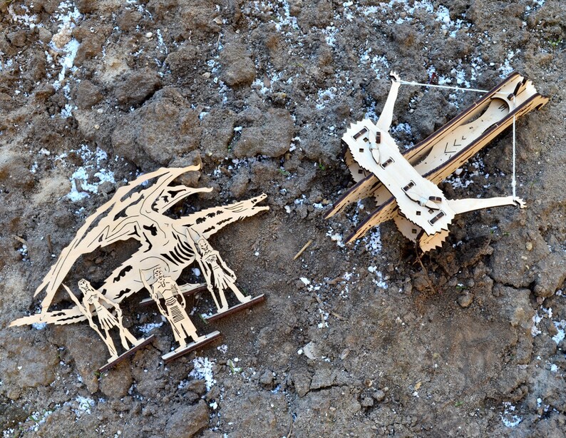 May include: A wooden dragon figurine with three figures standing in front of it. The dragon is facing a wooden catapult with a string attached to it.