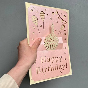 Side angle handheld view of the dimensional 3D cupcake pop-up card template, illustrating precise laser cutting work for the intricate pop-up details of the cupcake and balloons in a pink paper design.