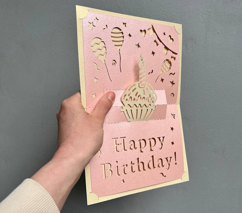 Side angle handheld view of the dimensional 3D cupcake pop-up card template, illustrating precise laser cutting work for the intricate pop-up details of the cupcake and balloons in a pink paper design.