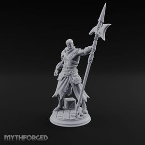 May include: A light gray unpainted resin miniature figure of a warrior standing on a circular cobblestone base. The figure wears layered armor and a long tunic, holding a tall polearm with a spear tip. A small crate sits at the feet.