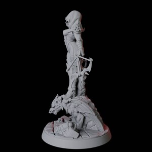 May include: A gray figurine of a hooded figure holding a crossbow, standing on a detailed base with a wolf-like creature. The figure is wearing armor and boots. The base is round and has intricate details.