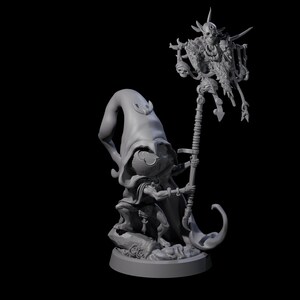 Può includere: Statuetta grigia di un personaggio fantasy con un grande cappello a punta e mantello, che tiene in mano un bastone sormontato da una figura scheletrica. Il personaggio è inginocchiato su una base rocciosa, con dettagli.