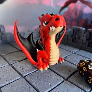Peut inclure: Figurine de dragon rouge et noir avec un ventre beige et des dents blanches. Le dragon a de grandes ailes noires et est posé sur une surface de pierre grise. Un dé noir et or est au premier plan.