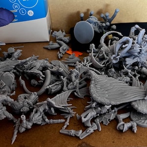 Può includere: Una collezione di figurine in miniatura grigie non verniciate, probabilmente per giochi da tavolo o modellismo. L'assortimento comprende varie creature, personaggi ed elementi del terreno, suggerendo un tema fantasy o fantascientifico. I pezzi sono sparsi su una superficie di legno.
