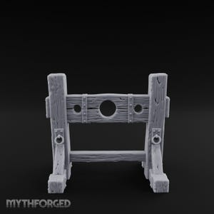 May include: A gray miniature pillory model featuring a textured wood grain finish, two vertical support posts, a horizontal beam with three circular openings, and two metal-look rings attached to the front. The text MYTHFORGED is visible.