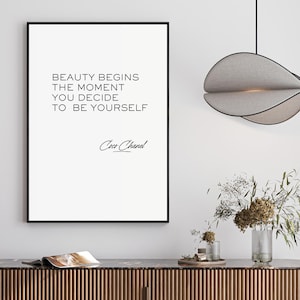 Peut inclure: Impression encadrée avec la citation « BEAUTY BEGINS THE MOMENT YOU DECIDE TO BE YOURSELF » de Coco Chanel. L'impression est au-dessus d'un meuble en bois, avec une lampe et des fleurs.