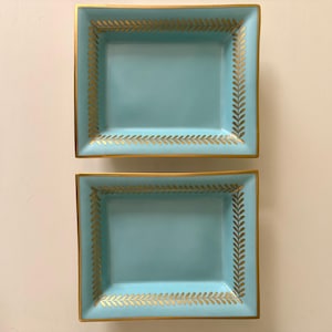 Peut inclure: Deux plateaux décoratifs rectangulaires bleu clair avec une bordure et des motifs dorés. Les plateaux ont une bordure dorée sur les bords et un motif de feuilles d'or sur les côtés.
