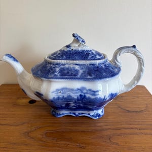 May include: A vintage blue and white porcelain teapot. The teapot has a landscape design, a curved spout, and a looped handle. The lid has a decorative finial. The design includes a blue and white landscape scene.