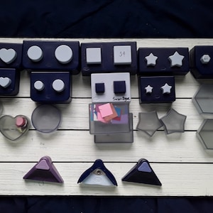 Puede incluir: Una colección de 18 punzones artesanales de diferentes formas, incluyendo corazones, círculos, estrellas, cuadrados y hexágonos. Los punzones están hechos de plástico y tienen una combinación de colores azul, gris y morado. Algunos de los punzones tienen cubiertas de plástico transparente.
